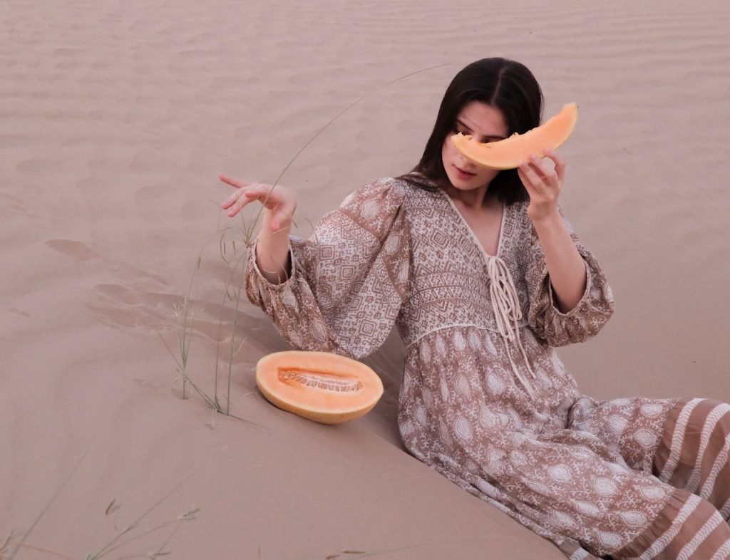 Woman sitting in Dubai desert holding up melon slice. A serene summer scene showcasing relaxation and natural beauty.