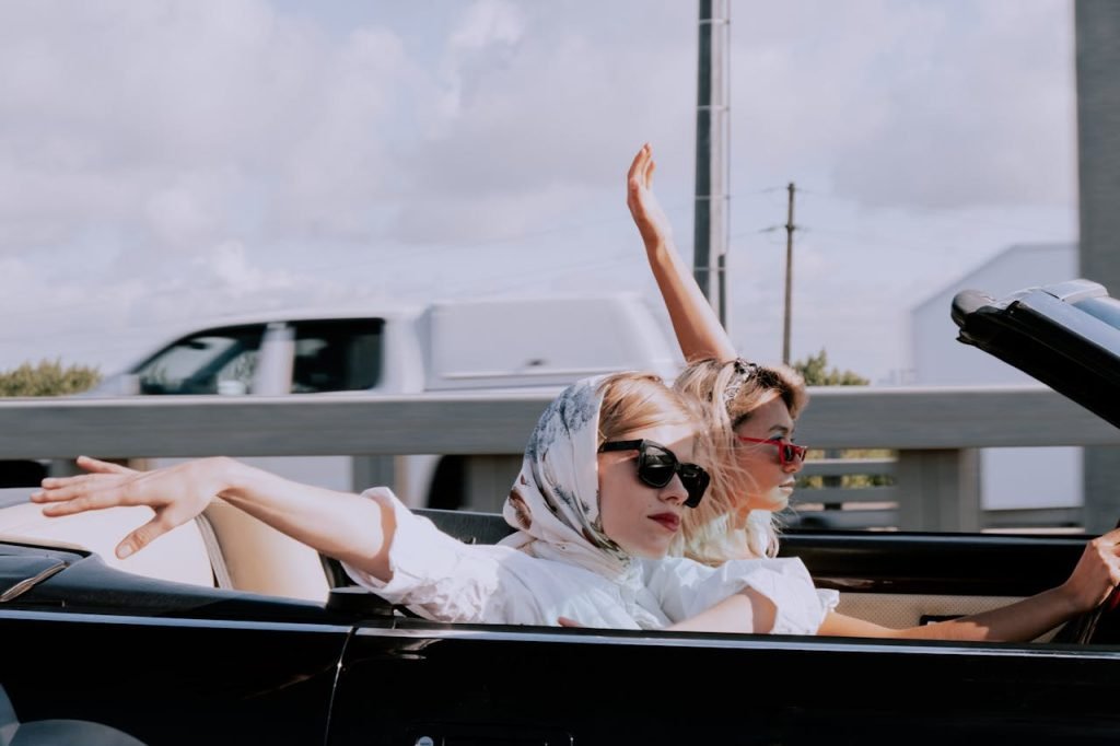 Two women in a convertible car, wearing sunglasses and headscarves, enjoying a drive with hands raised